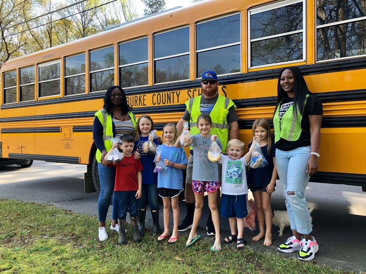 Kids holding lunches standing in front of school bus with 3 school bus drivers in safety vests. 